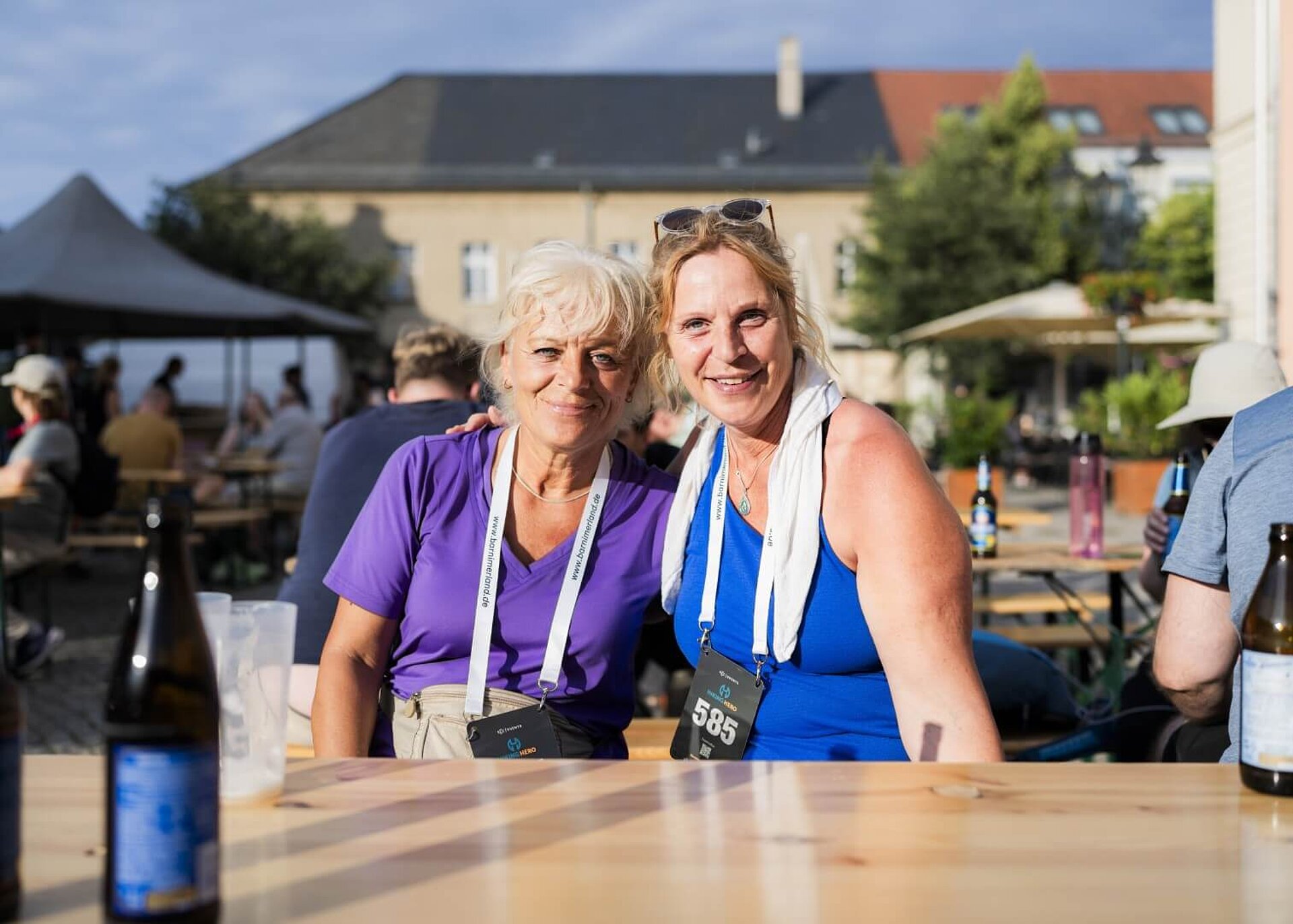 Zwei lächelnde Teilnehmer:innen erholen sich auf einer Bierzeltgarnitur mit Getränken.