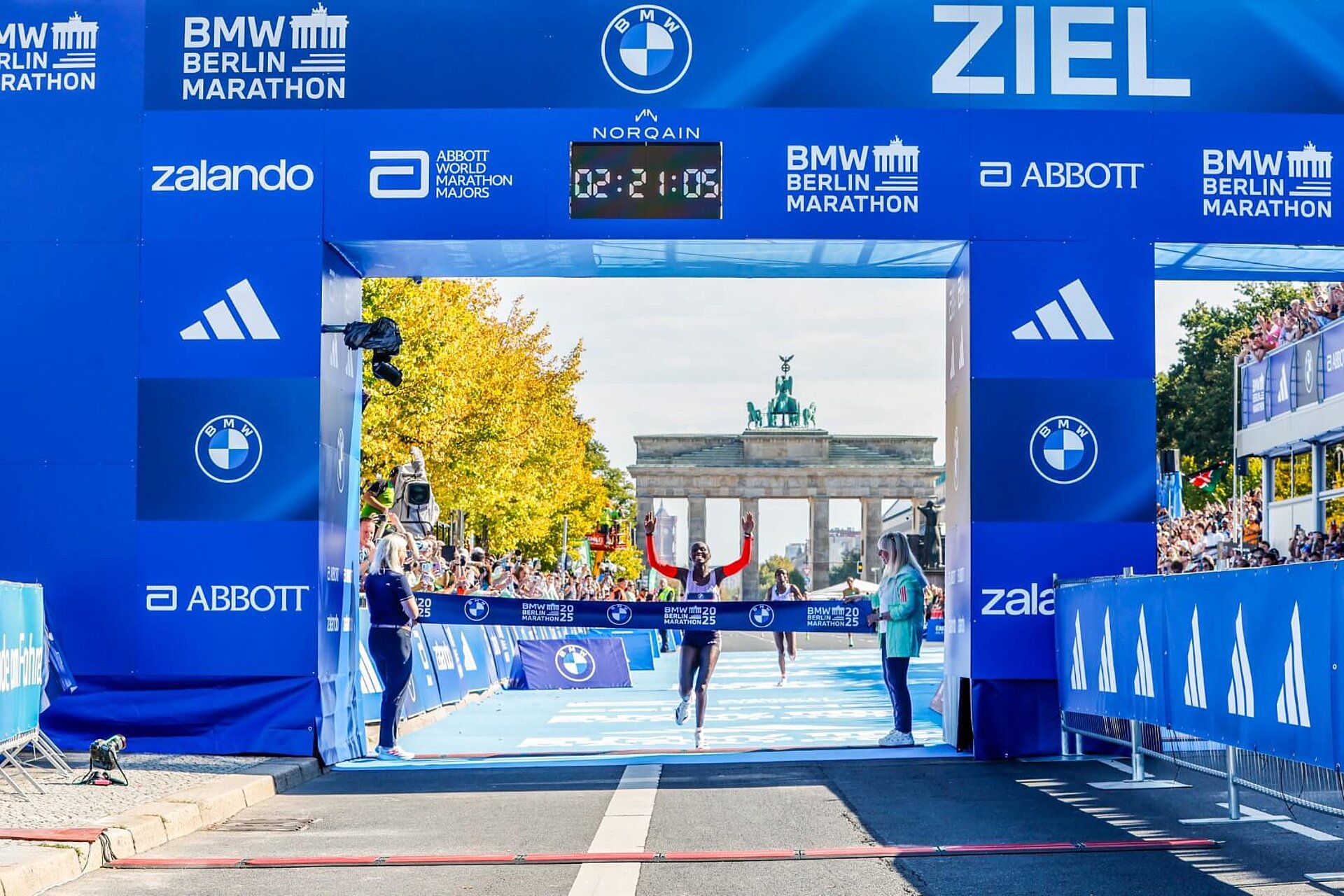 Rosemary Wanjiru durchläuft das Zielband des BERLIN-MARATHON 2025. Über ihr zeigt die Uhr auf dem Zieltor 02:21:05 an.