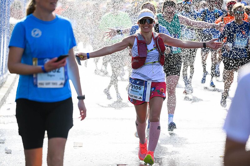 Runner at the BERLIN-MARATHON wearing a hat and sunglasses smiles as she runs through a shower of water..