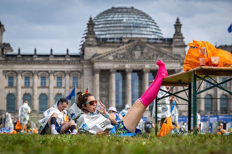 A marathon finisher is sitting in front of the Reichstag, drinking something and resting her legs.