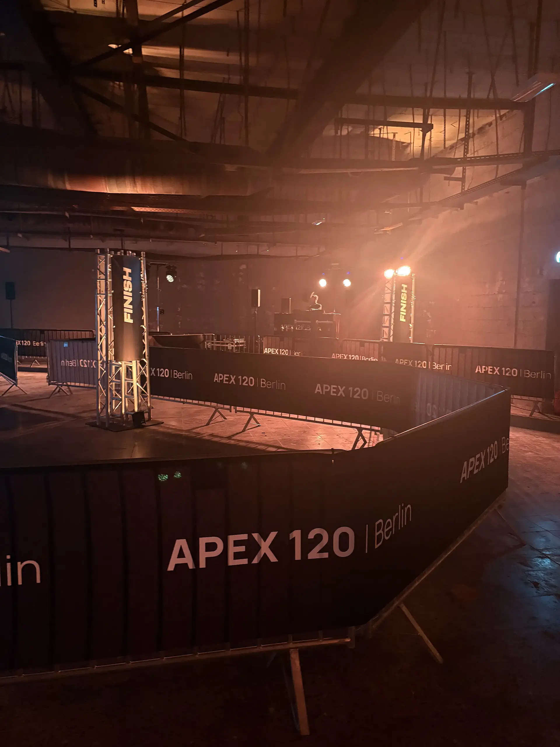 Halle in warmem, dramatischem Licht mit Streckenbegrenzungen auf denen "APEX 120 Berlin" steht. Im Hintergrund ein DJ hinter seinem Pult.