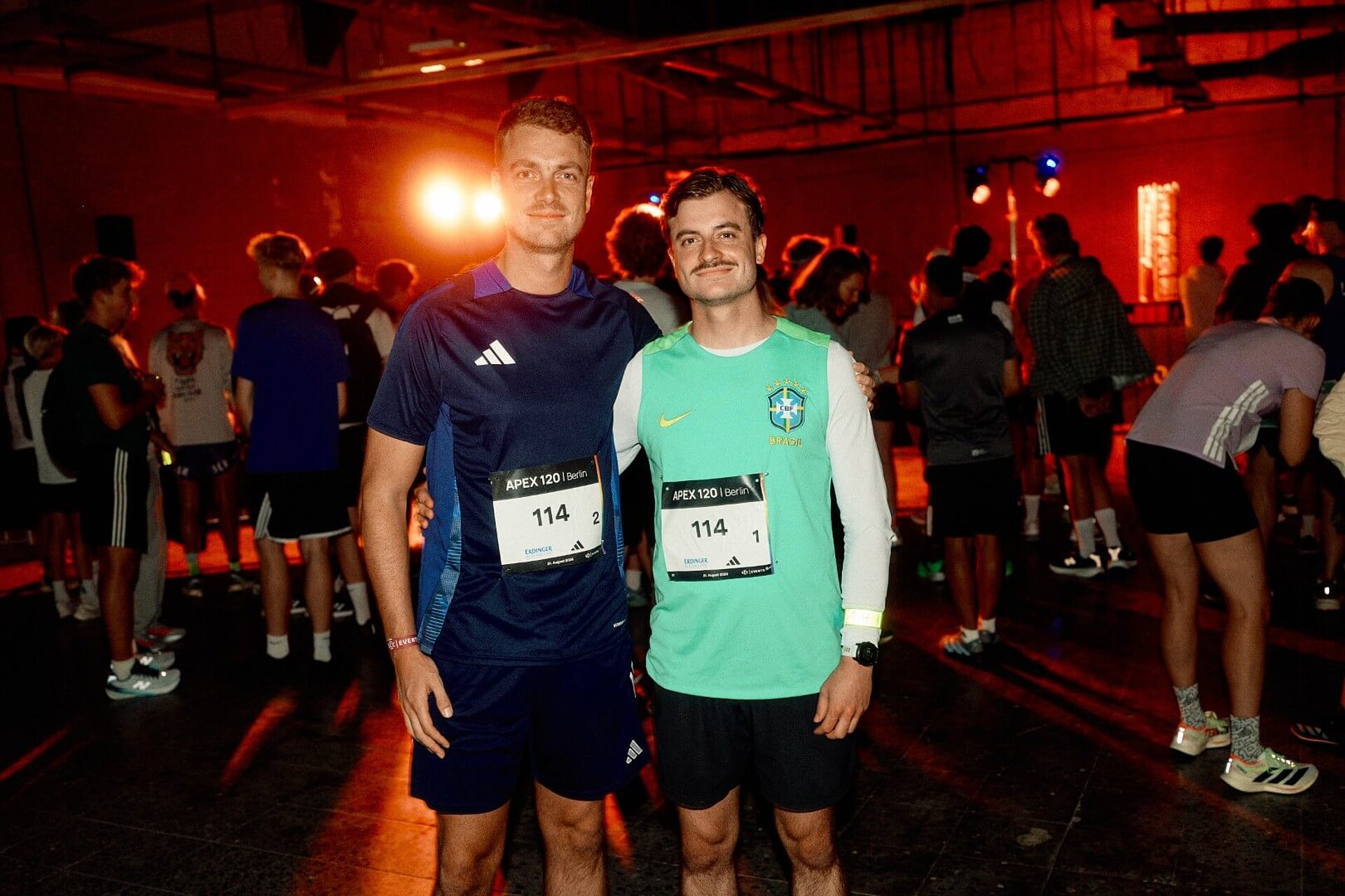 Jakob und Micha, Arm in Arm, lächeln in einer rot erleuchteten Halle in die Kamera.