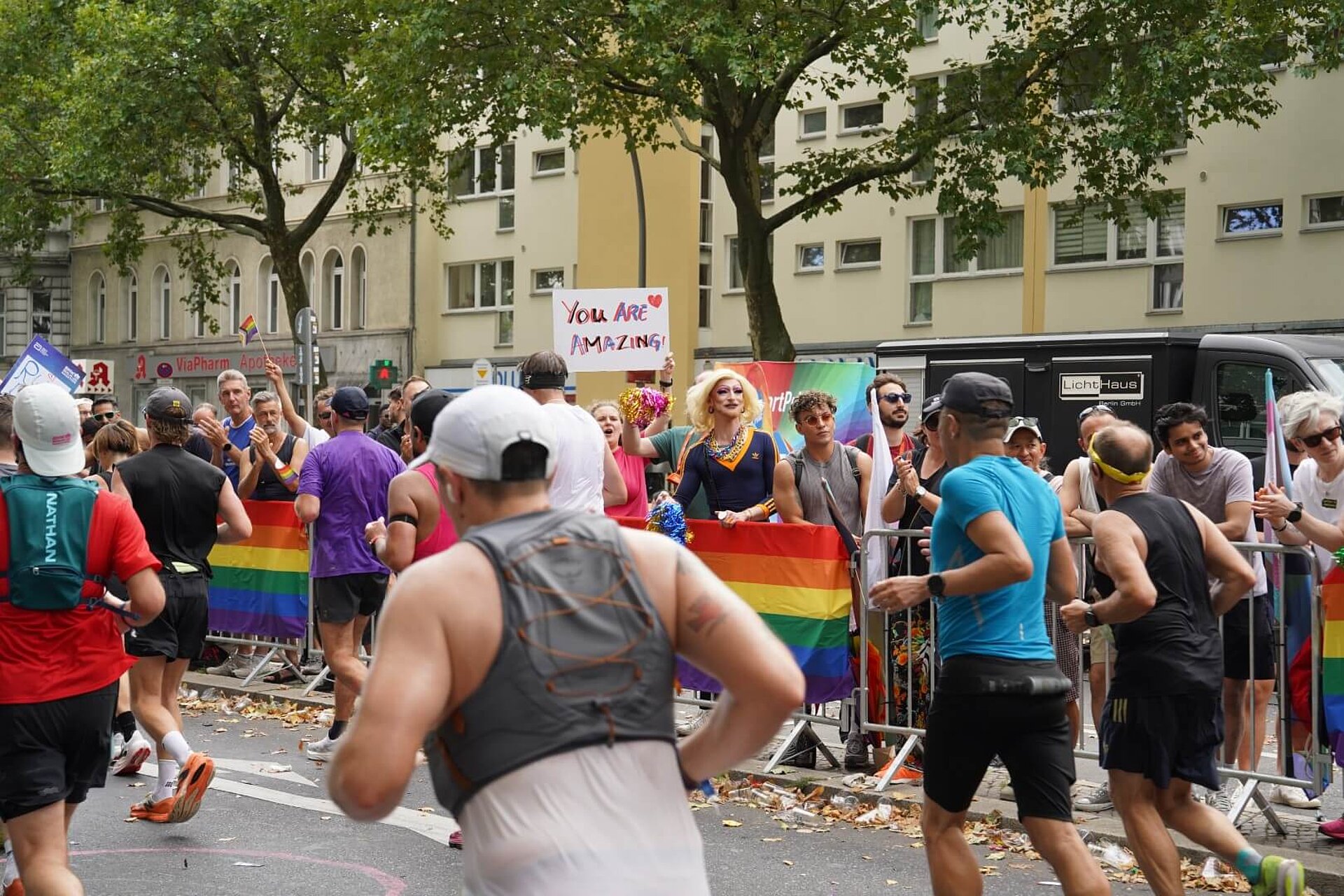 Queer Cheer mit Regenbogenflaggen an der Streckenbegrenzung und einer Person in Drag.